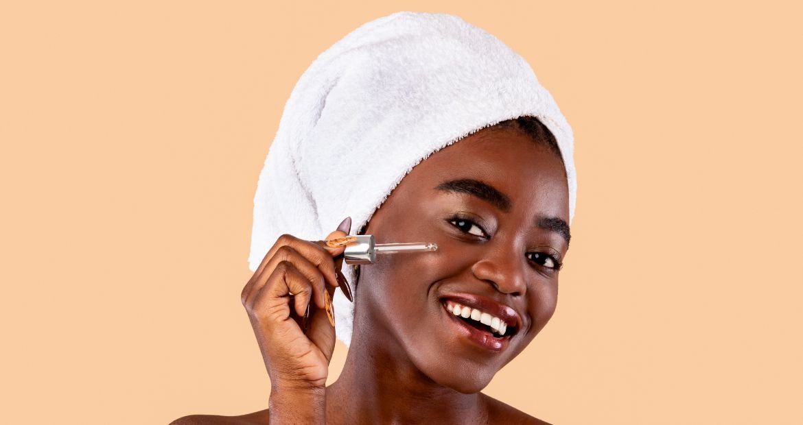Mulher com pele negra sorridente passando conta gotas no rosto e com toalha branca no cabelo.